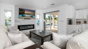 Ein modernes Wohnzimmer mit Kamin, einem an einer weißen Wand montierten Fernseher und Blick auf Pool und Außenbereich.