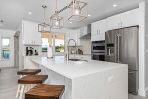 A modern kitchen with white cabinetry, a large island, stainless steel appliances, and wooden stools. - Bonita Lake Home - Luxury taken to the next level (Bonita Springs)