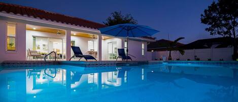Piscina con sillas de descanso y sombrillas, rodeada por un edificio con tejado de baldosas y puertas de cristal.