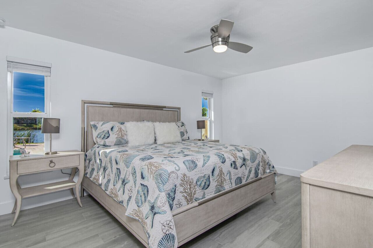 Un dormitorio con cama, mesitas de noche, ventilador de techo y ventanas.