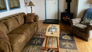 A cozy living room with a brown sofa, patterned armchair, and a small wooden table. A deer head is mounted on the wall, and a wood-burning stove is visible. - Upper Moose Caboose (Homer)