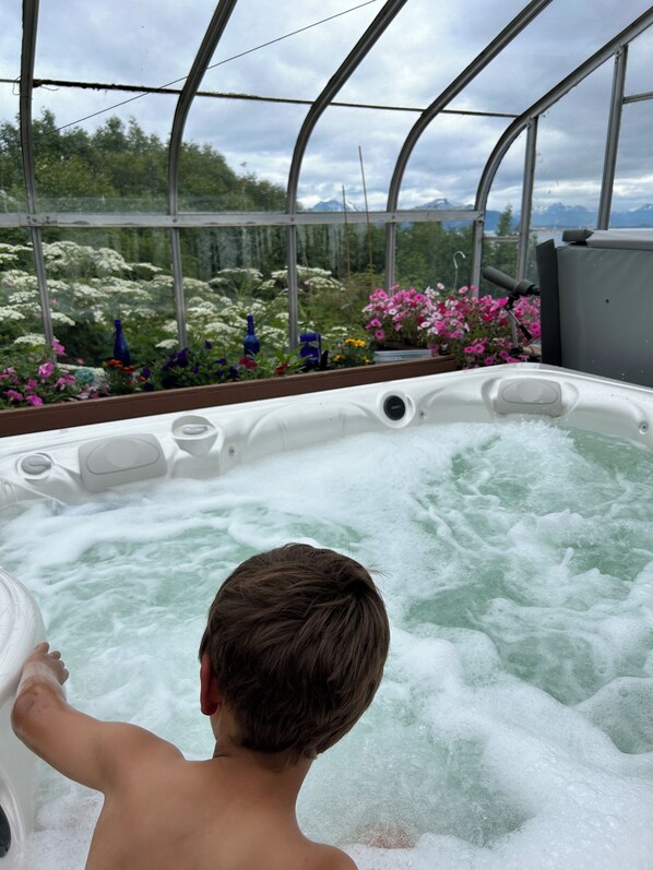 A person in a hot tub with a view of a greenhouse filled with flowers. - Upper Moose Caboose (Homer)