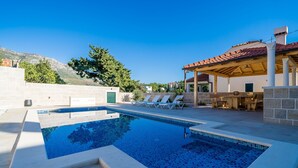 Una piscina con sillas de descanso, un porche cubierto y montañas al fondo.