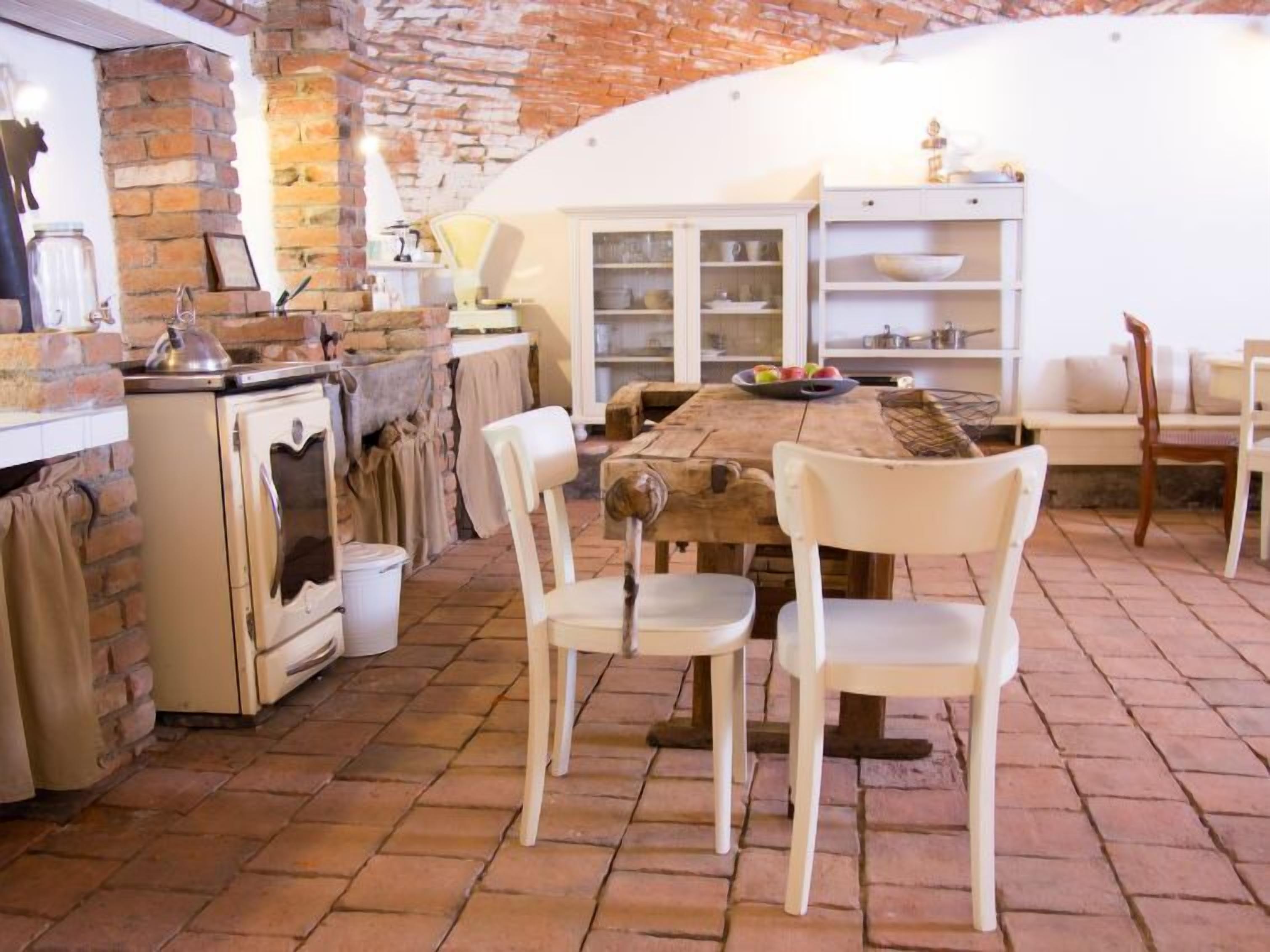 Eine rustikale Küche mit Backsteinwand, Holztisch, weißen Stühlen und einem weißen Schrank mit Glastüren.