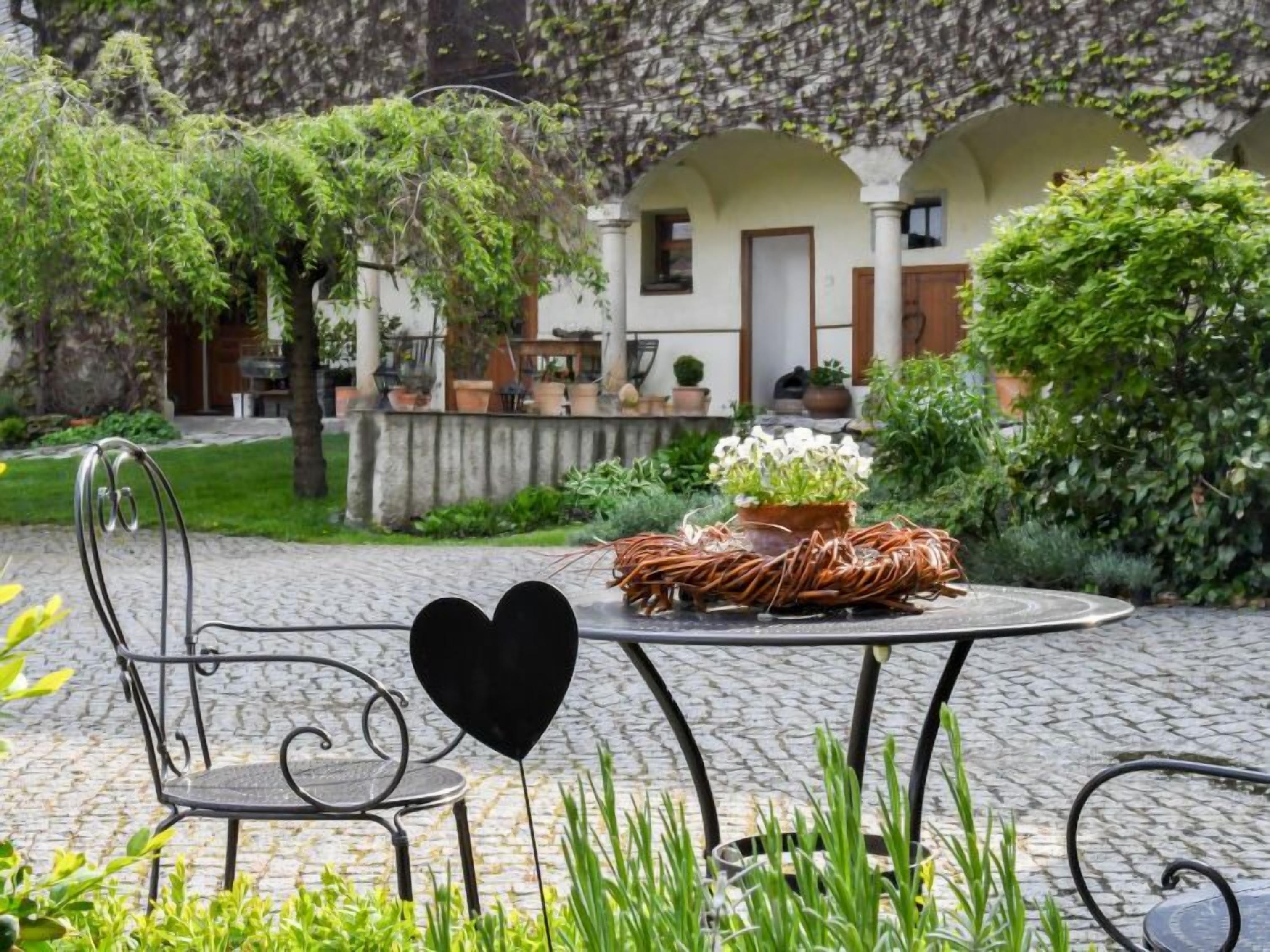 A garden patio with a metal table and chair, surrounded by greenery and a building with arches in the background.