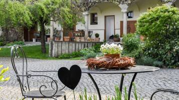 A garden patio with a metal table and chair, surrounded by greenery and a building with arches in the background.