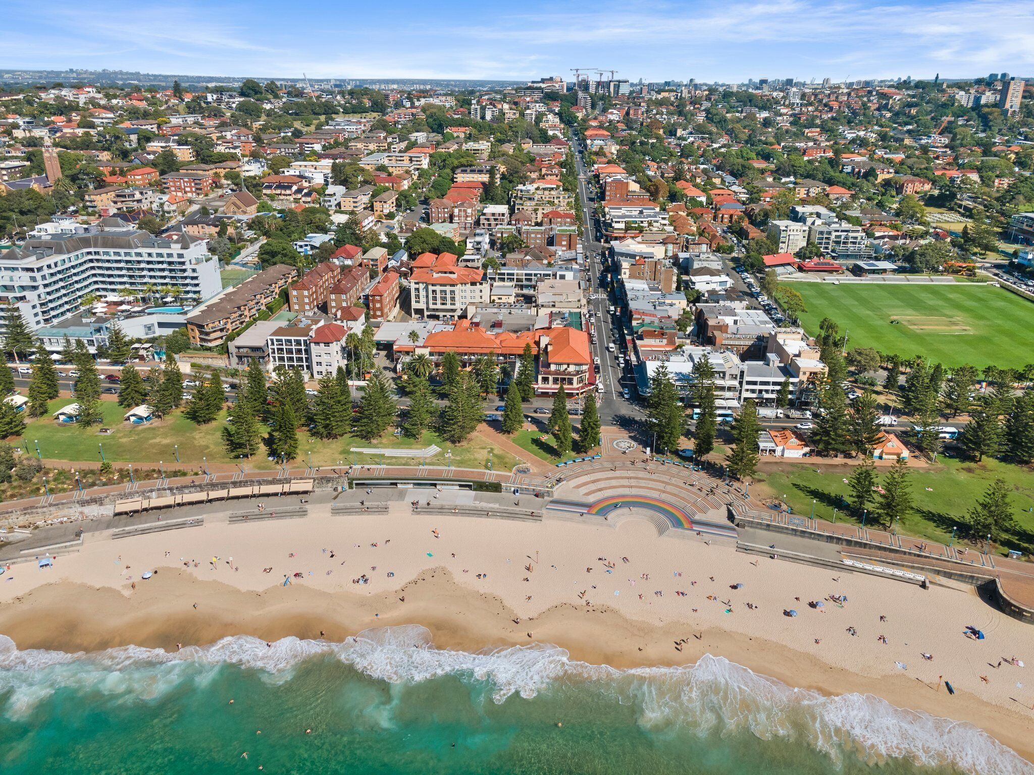 Luchtbeeld van een kuststad met een breed strand, woonwijken en een opvallende weg die door het centrum loopt.