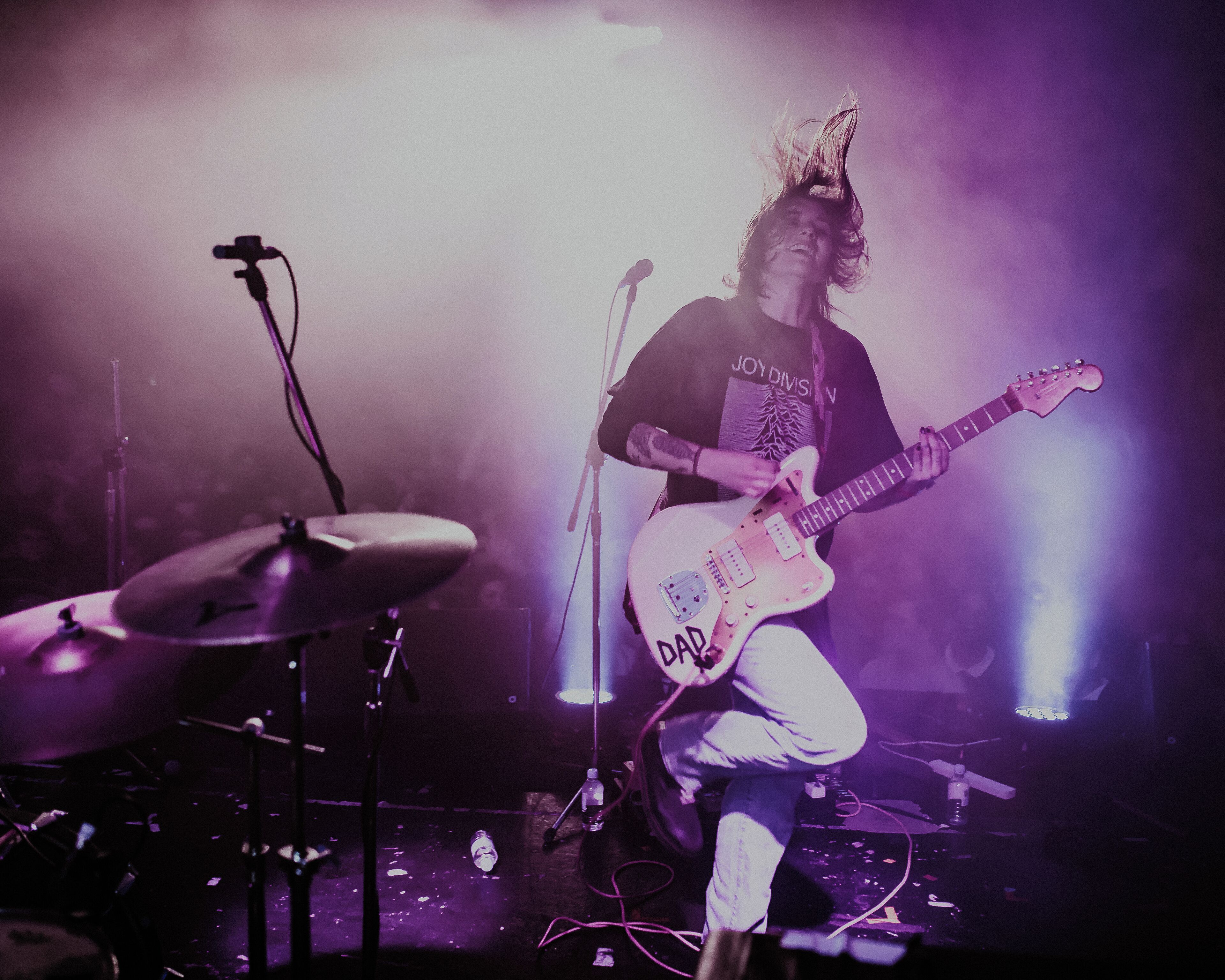 A musician performing on stage with a drum set and a guitar, surrounded by stage lighting.