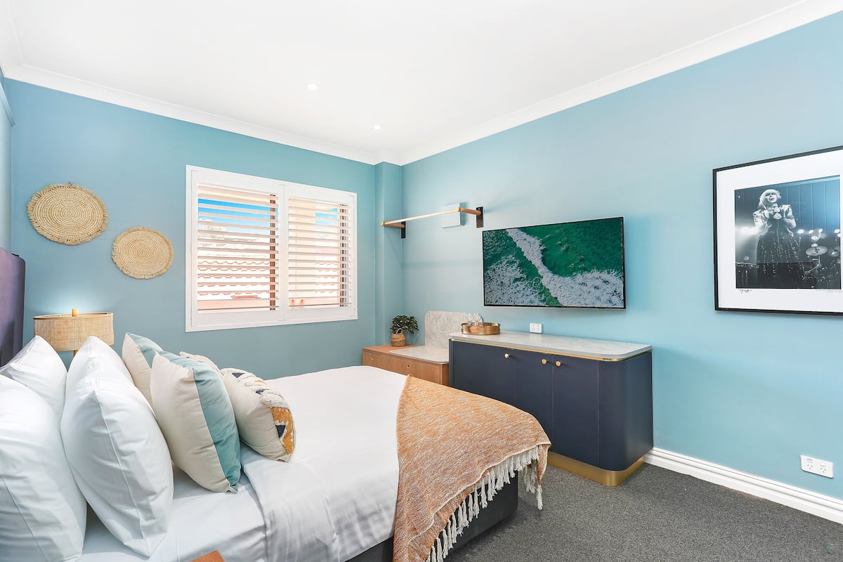 Une chambre à coucher comprenant un lit, une table de chevet, une commode et une grande fenêtre avec des stores.