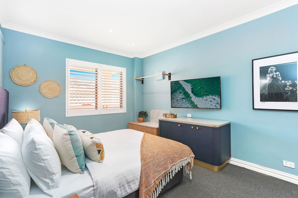 Une chambre à coucher comprenant un lit, une commode, un téléviseur et une fenêtre avec des stores.