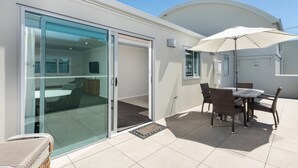 A modern house with a glass door leading to a balcony, a dining area with a table and chairs, and a white umbrella. - Modern Marine Parade Apartment (Bay of Plenty)