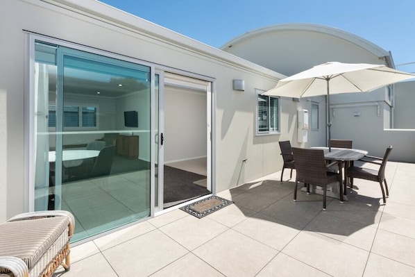 A modern house with a glass door leading to a balcony, a dining area with a table and chairs, and a white umbrella. - Modern Marine Parade Apartment (Bay of Plenty)