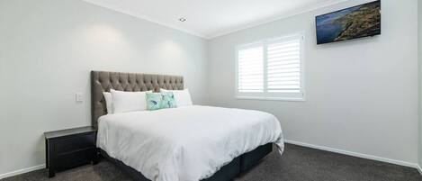 A bedroom with a tufted headboard bed, a nightstand, a window with blinds, and a wall-mounted TV.