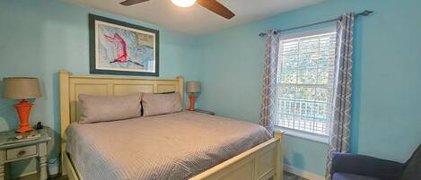 Quarto com uma cama, duas mesas de cabeceira, um ventilador de teto, uma janela com cortinas e um quadro na parede.