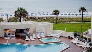 A pool area with multiple pools and lounge chairs, a building with a covered seating area, and a view of the beach with umbrellas.