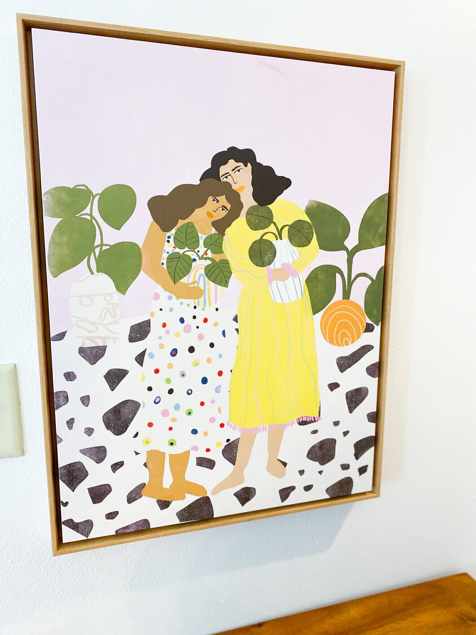 Une œuvre d’art encadrée représentant deux femmes entourées de plantes, le tout sur fond de mur blanc.