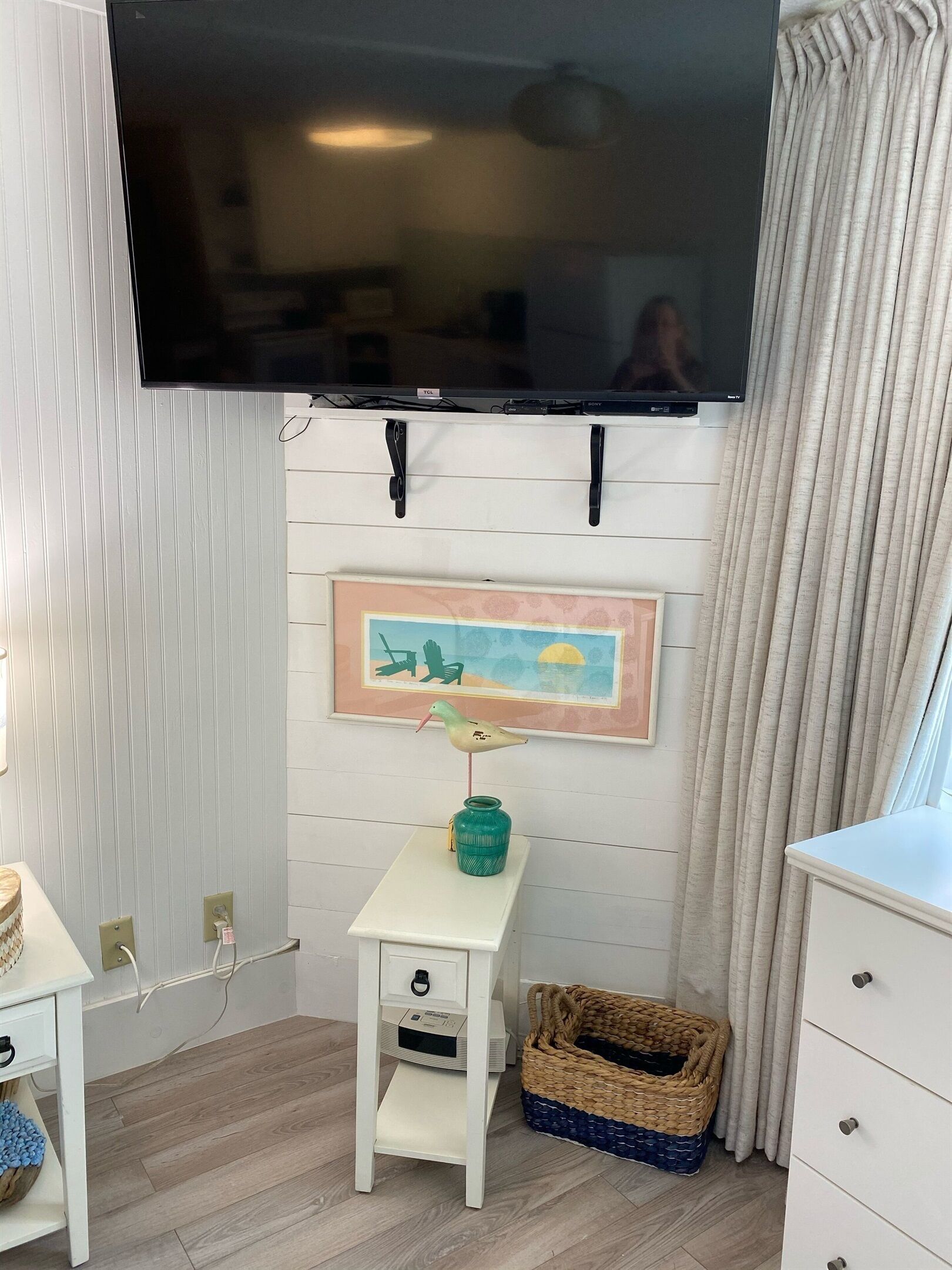 Een kamer met een wandgemonteerde flatscreen-tv, een klein wit tafeltje met een vaas en een lijstfoto, een rieten mand en een witte dressoir.
