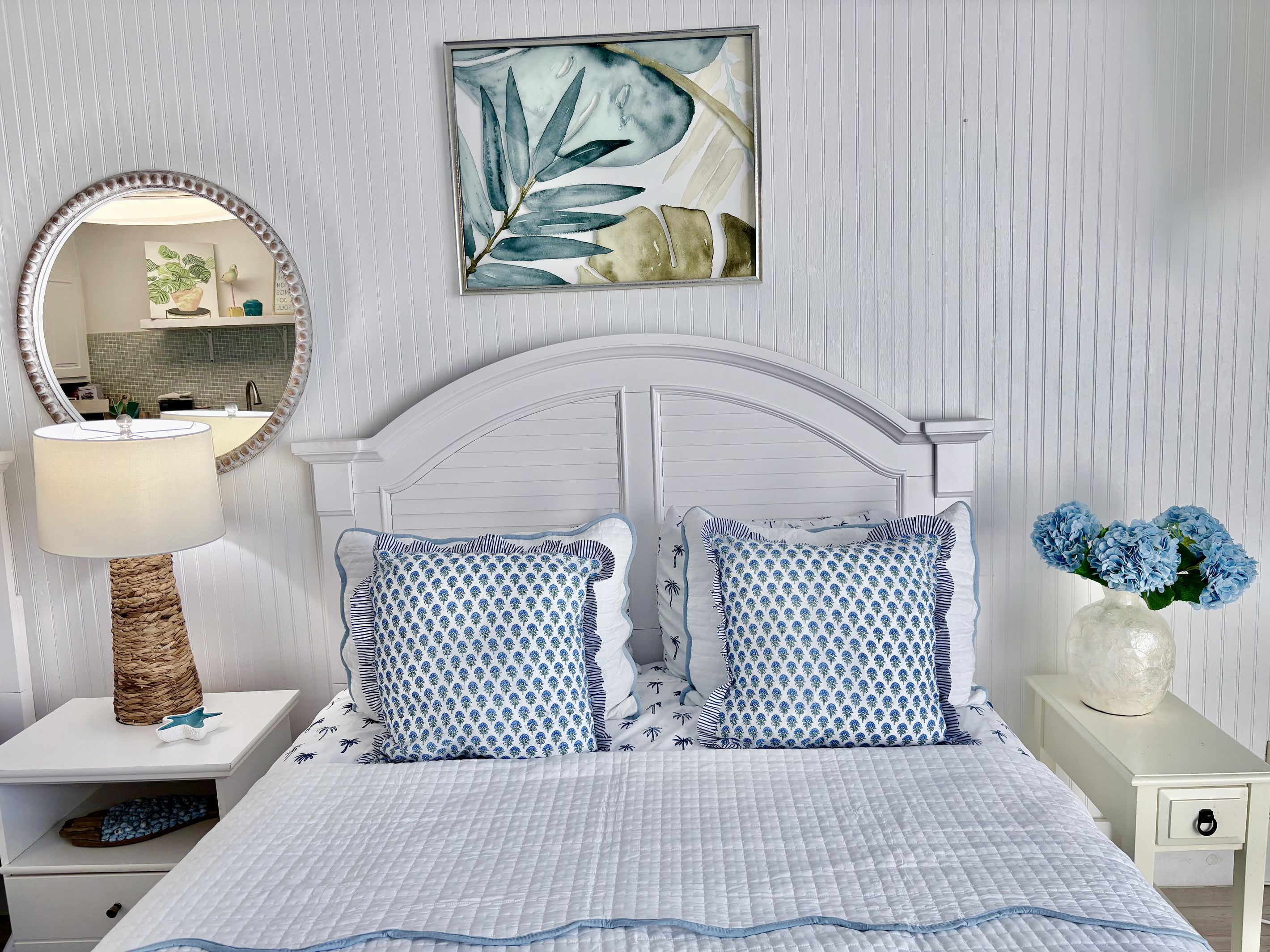A bedroom with a bed, pillows, a bedside table, a lamp, a mirror, and a wall art piece.