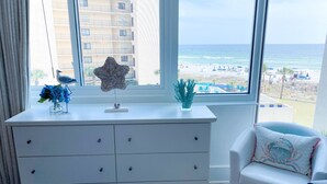 A room with a white dresser, a chair, and a view of the beach and ocean through large windows. - 4th floor beachfront studio with two queens (Panama City Beach)