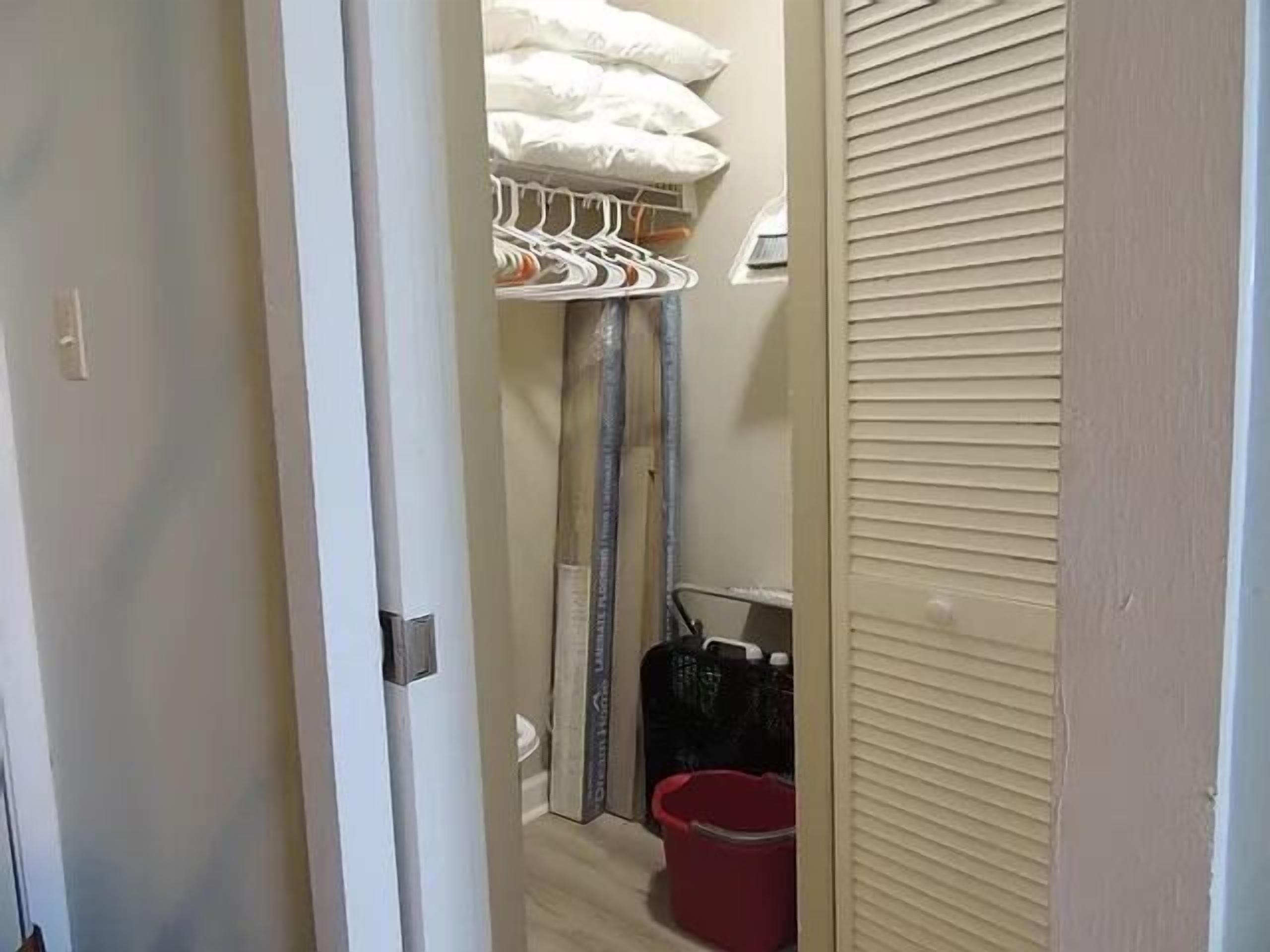 A narrow hallway with a closet containing hanging clothes and a red bucket.