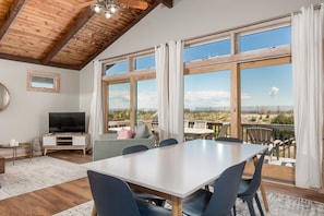 A spacious living area with a large table and chairs, a television, and a balcony with a view of the landscape.