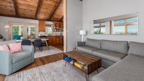 A spacious living room with a wooden ceiling, a grey sofa, a coffee table, and a dining area with a kitchen in the background. - Bayfront on Broadkill Beach! 1810 S Bayshore (Milton)