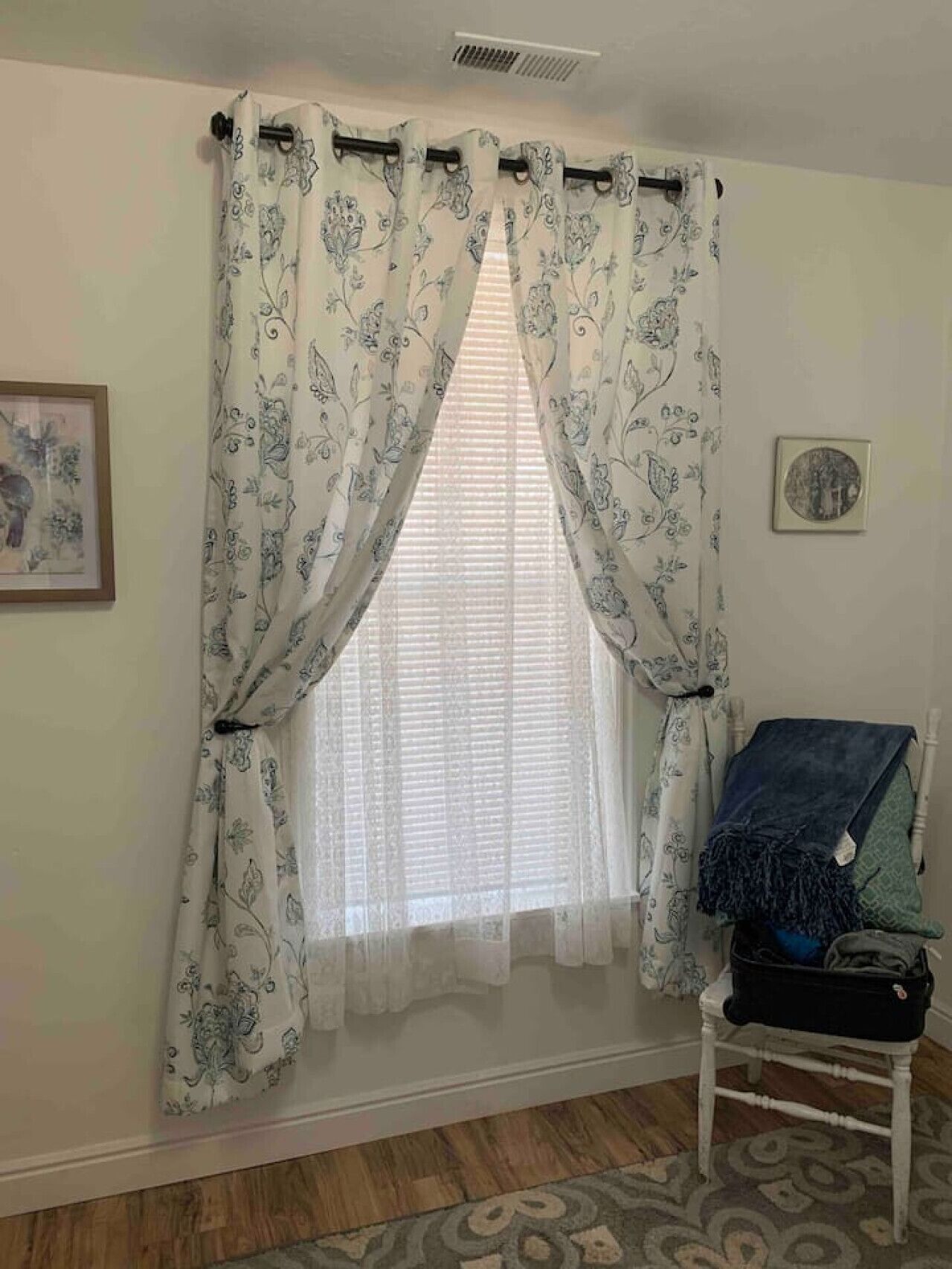 A room with a window covered by floral-patterned curtains, a framed picture on the wall, and a chair with a blanket.