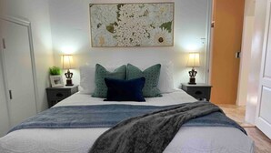 A neatly made bed with a white duvet, two pillows, and a grey throw blanket. There are two bedside tables with lamps and a framed picture on one. A large floral artwork hangs above the bed. - Jailer`s Cottage | 130-Year-Old Gem | Sleeps 12 (Spring City)