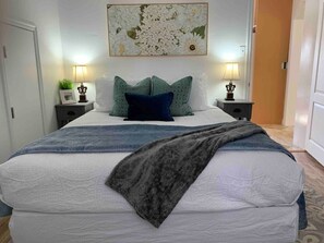 A neatly made bed with a white duvet, two pillows, and a grey throw blanket. There are two bedside tables with lamps and a framed picture on one. A large floral artwork hangs above the bed. - Jailer`s Cottage | 130-Year-Old Gem | Sleeps 12 (Spring City)