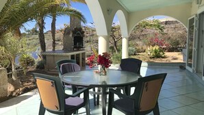 Eine überdachte Terrasse mit rundem Tisch und Stühlen, einem Kamin aus Stein und Blick auf eine Wüstenlandschaft mit Palmen und blühenden Pflanzen.