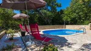 Una piscina con un patio de piedra, dos sillas reclinables rojas y una sombrilla grande.