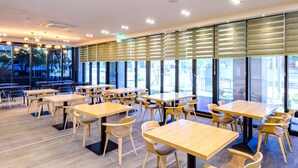 Un restaurant moderne avec des tables et des chaises en bois, de grandes fenêtres équipées de stores et un intérieur lumineux.