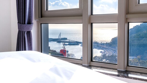 Une chambre d’hôtel dotée d’une grande fenêtre donnant sur une zone côtière, avec vue sur un pont et un bâtiment rouge.