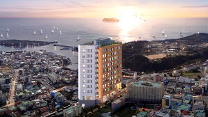 Un paysage urbain côtier avec un grand hôtel doté de nombreux balcons, un port de plaisance avec des voiliers et un coucher de soleil sur l’eau.