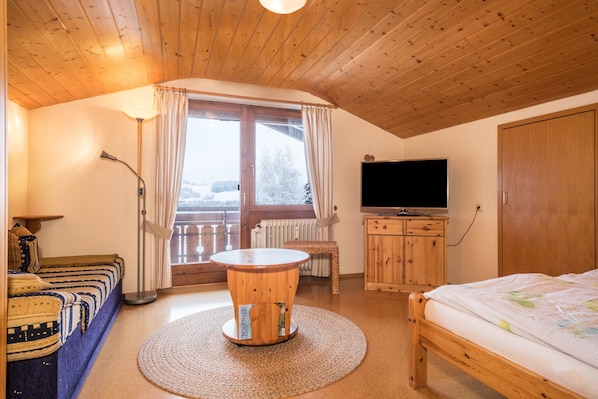 Una stanza con soffitto in legno, un letto, una televisione, un divano, un tavolo rotondo in legno e una finestra con tende.
