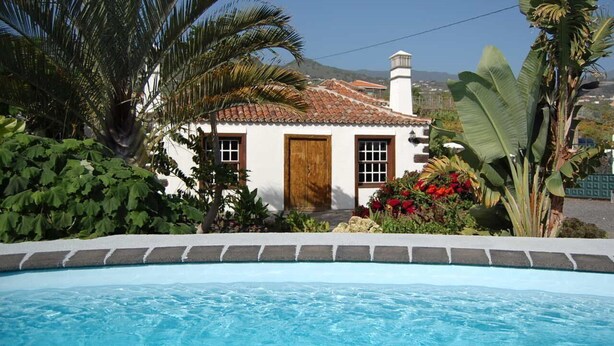 Un edificio blanco con techo de tejas, puerta de madera y una piscina al frente.