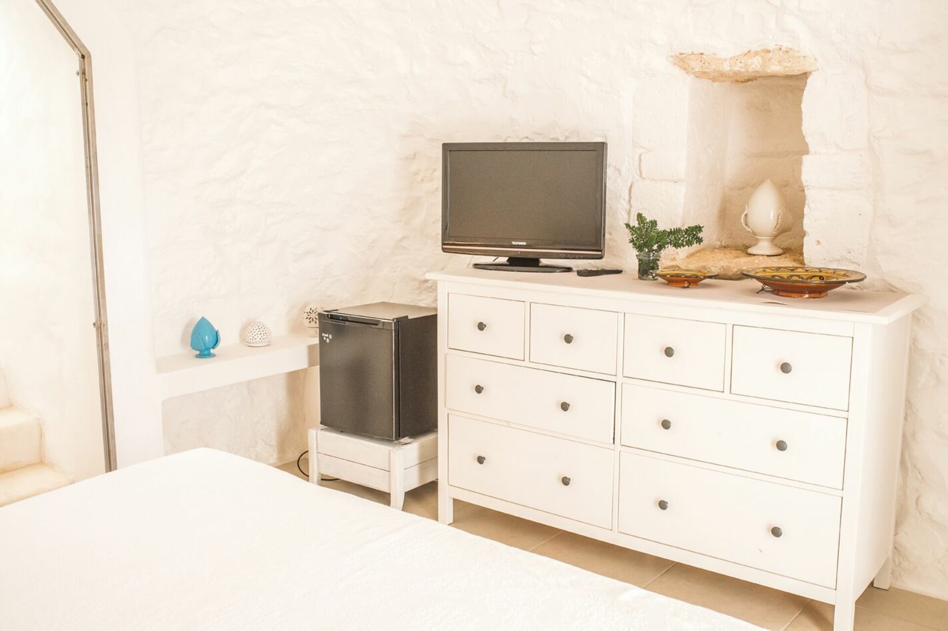 A room with a white dresser, a television on top, a small refrigerator, and decorative items.