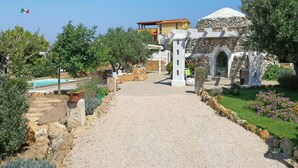 Un chemin mène à un bâtiment à la façade en pierre et surmonté d’un dôme, entouré de verdure et d’une piscine.