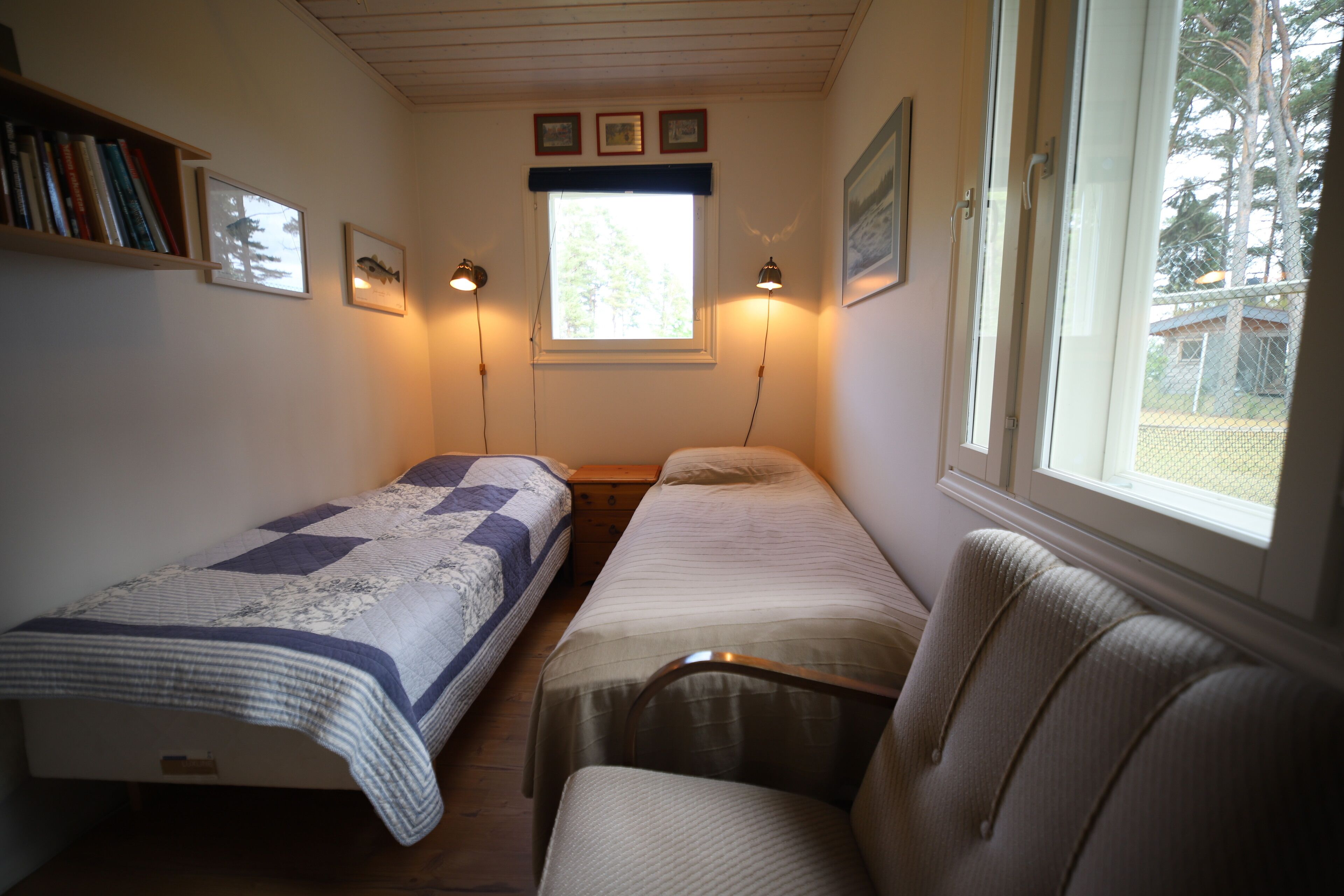 Una habitación con dos camas individuales, una silla, una estantería y una ventana con vistas a árboles.