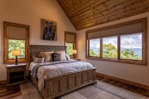A bedroom with a wooden bed, two nightstands, a large window with a view, and a painting on the wall.