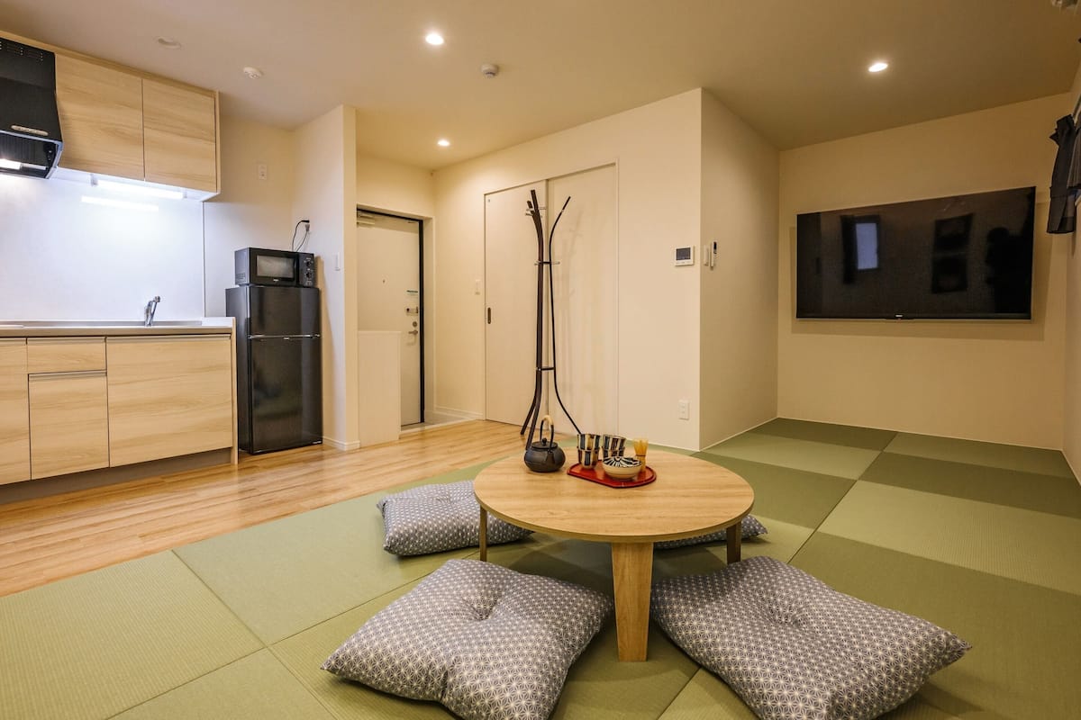 A modern kitchen with wooden cabinets, a black refrigerator, and a microwave. There is a dining area with a round table and cushions.