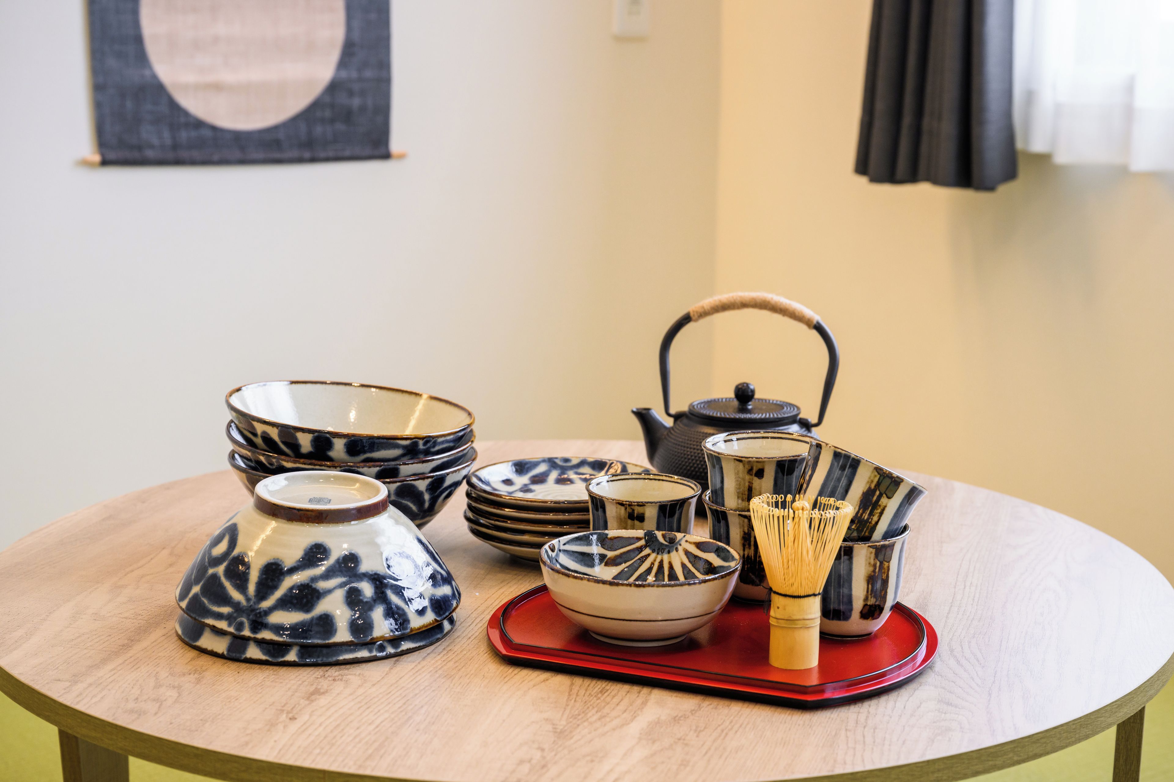 A round wooden table with a variety of ceramic tea ware, including bowls, a teapot, and a whisk set, arranged on a red tray.