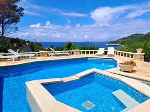Piscina com vista para o mar, cercada por espreguiçadeiras e um balustrade de pedra.