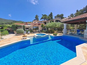 Ein Poolbereich mit klarem, blauem Wasser, Steinstufen, die zum Pool führen, und ein Haus mit Steinbauweise und einem Ziegeldach im Hintergrund.