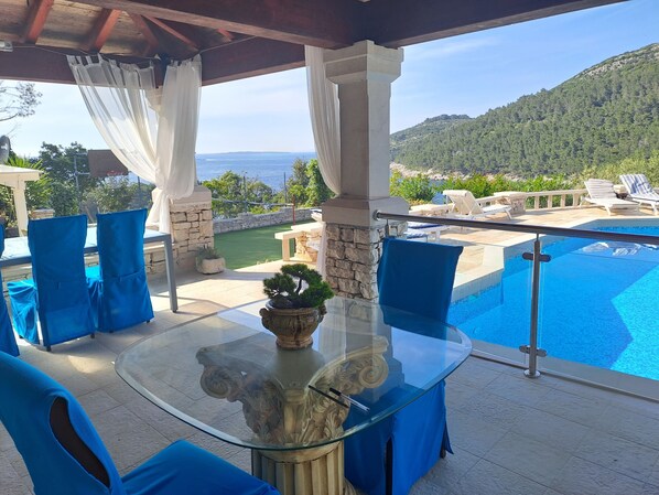 Espaço ao ar livre com mesa de vidro, cadeiras azuis e vista para uma piscina e montanhas.