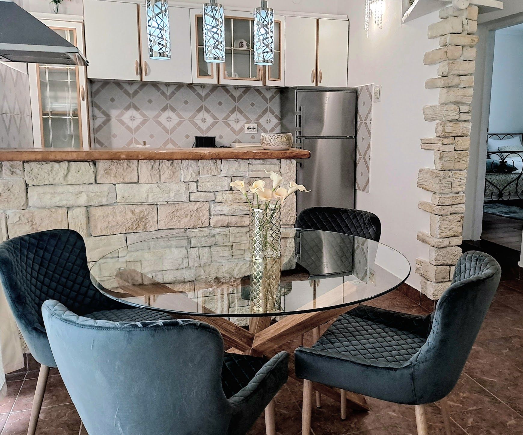 A modern kitchen with a stone wall, a stainless steel refrigerator, and a glass dining table with two chairs.