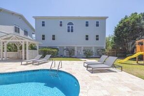 Une piscine dotée de chaises longues et d’un toboggan, entourée d’une cour clôturée et d’une grande maison.