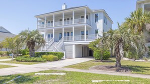 A two-story beachfront house with a balcony, palm trees, and a clear sky.