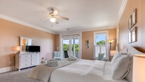 A bedroom with a large bed, a ceiling fan, a dresser, a TV, and a window with a view of greenery.