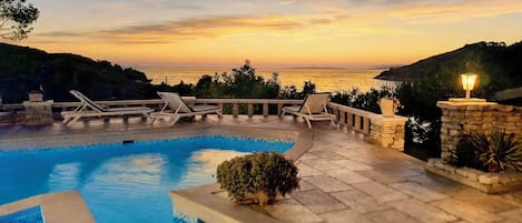 A swimming pool with lounge chairs and a view of the sea at sunset.